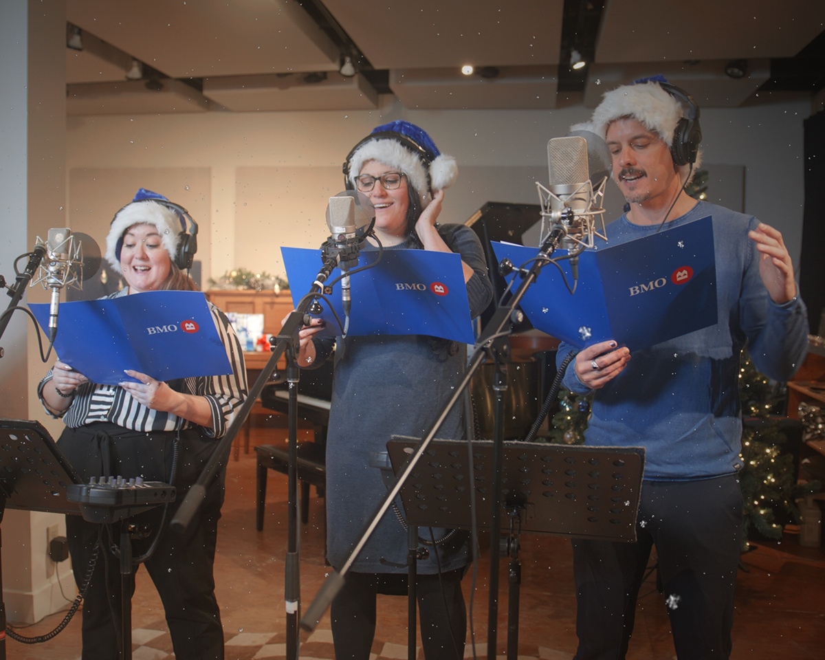 Trois chansonniers dans un studio d'enregistrement qui chantent une chanson des Fêtes. Ils portent des chapeaux de Père Noël bleus et tiennent des pochettes bleues avec le logo BMO.